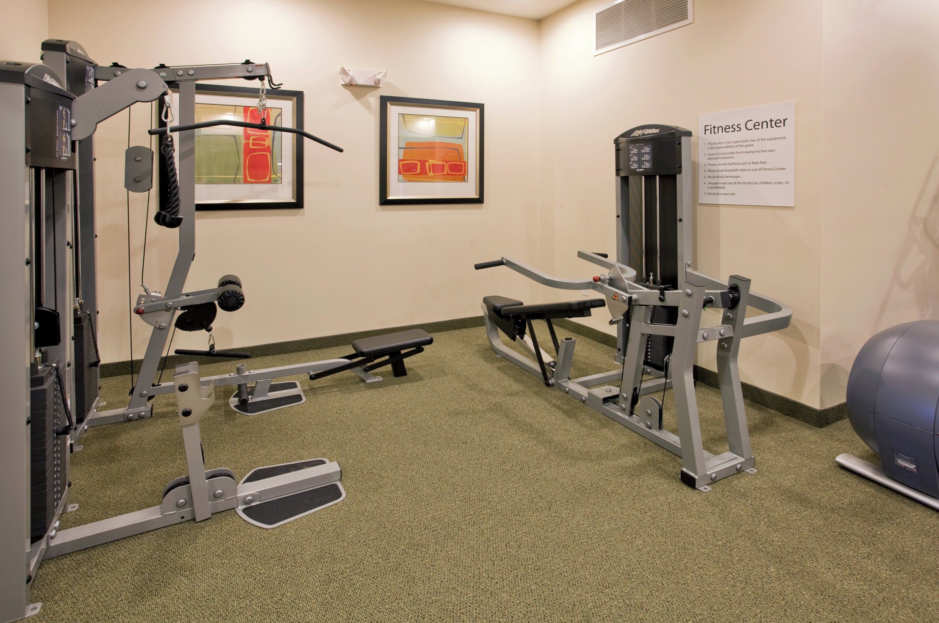 A fitness center with exercise equipment, including a leg press machine, a chest press machine, and a seated row machine. There are two framed artworks on the wall and a sign indicating the fitness center's rules.
