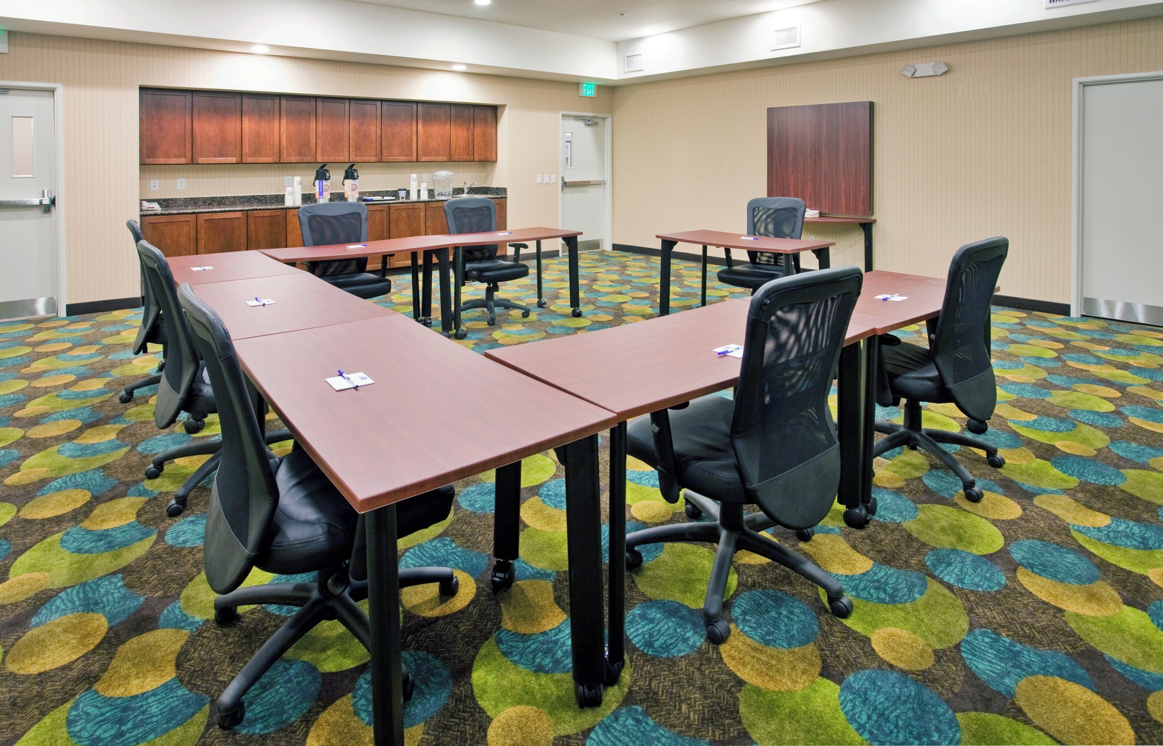 A conference room with a long table, chairs, a kitchenette, and a patterned carpet.