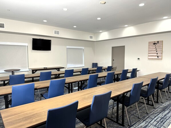 Un salón de conferencias con largas mesas, sillas azules, una pizarra, una pantalla de proyector y una puerta.