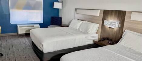 A hotel room with two beds, a large window with blinds, a bedside table with a lamp, and a wall-mounted light fixture.