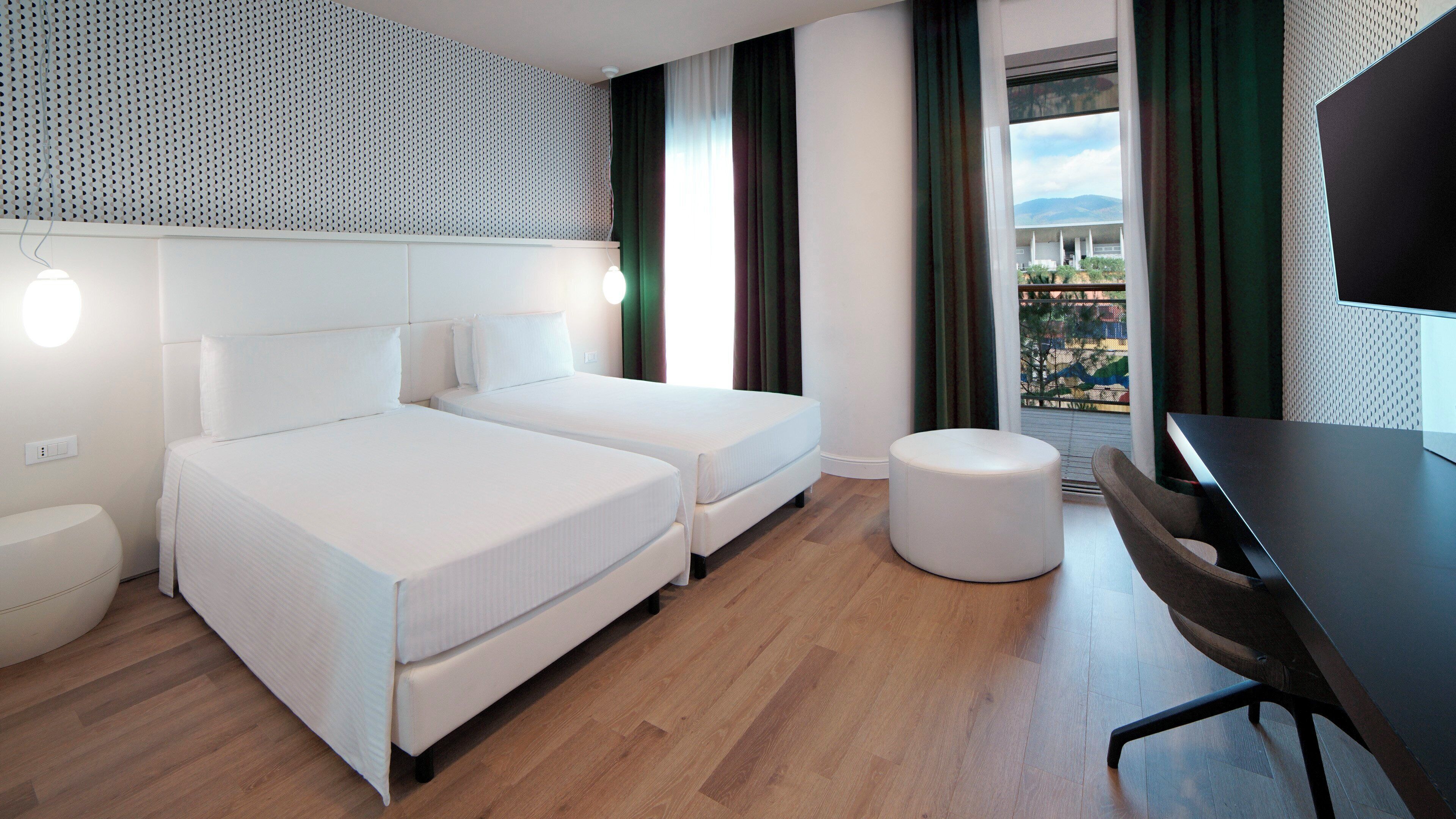 Una habitación de hotel moderna con dos camas, un otoman redondo, un escritorio y un amplio ventanal con vistas.