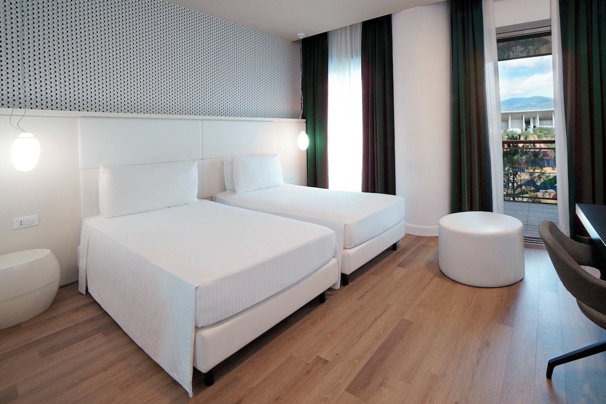 Ein modernes Hotelzimmer mit zwei Betten, einem runden Hocker, einem Schreibtisch und einem großen Fenster mit Aussicht.
