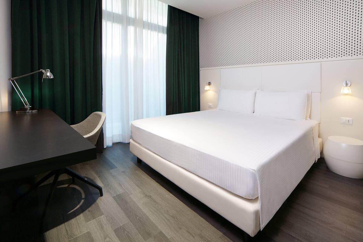A hotel room with a large bed, a desk, a chair, and a small bedside table.