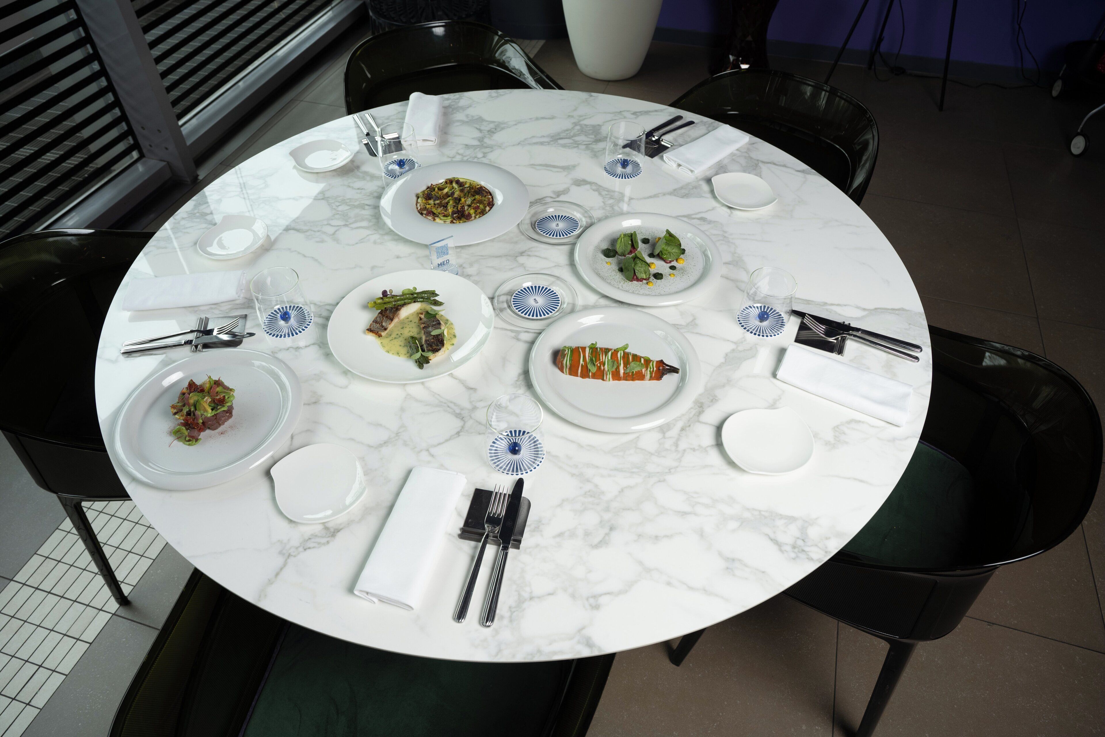 A round marble table with multiple plates of food, cutlery, and glasses.
