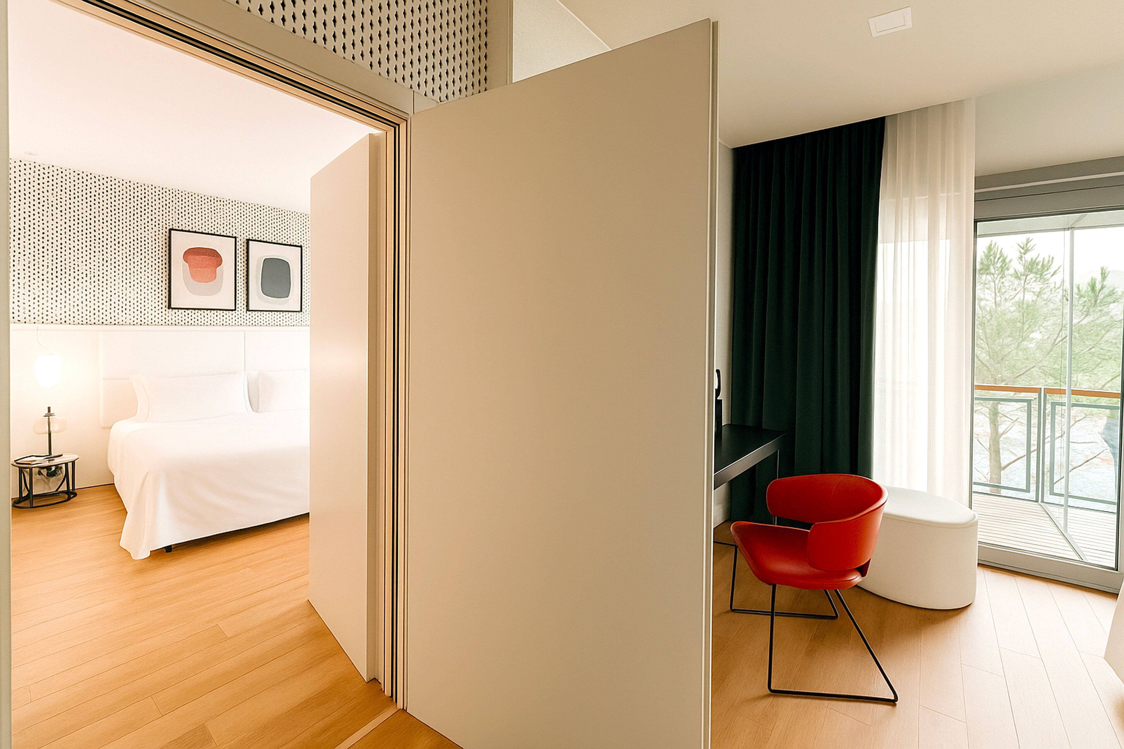 A modern hotel room with a bed, a red chair, a large window with curtains, and a wall with two framed pictures.