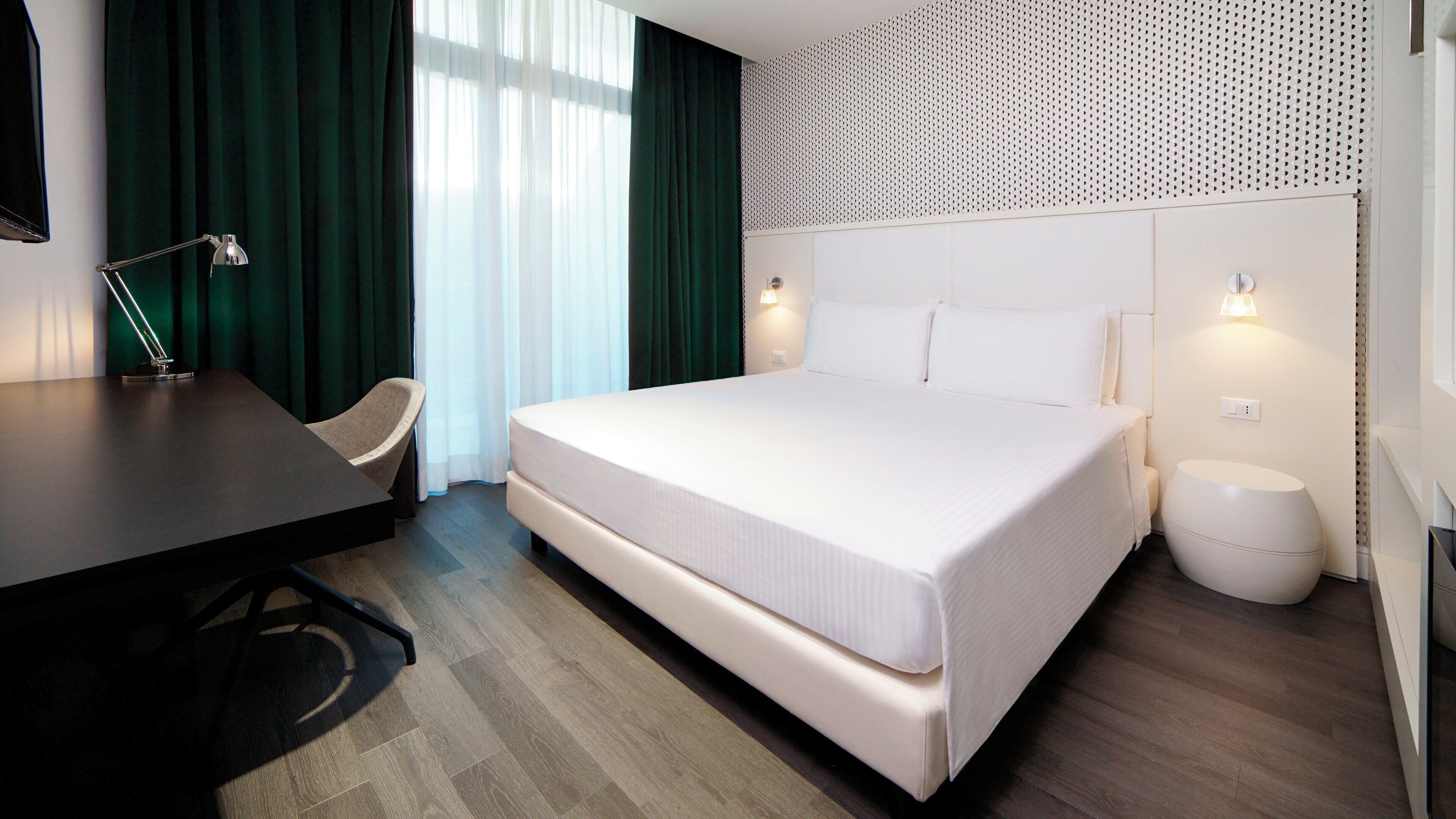 A modern hotel room with a bed, bedside table, chair, and a desk.
