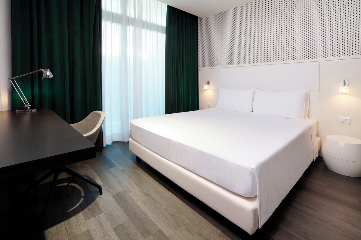 Ein Hotelzimmer mit einem großen Bett, einem Schreibtisch, einem Stuhl und einem kleinen Nachttisch.