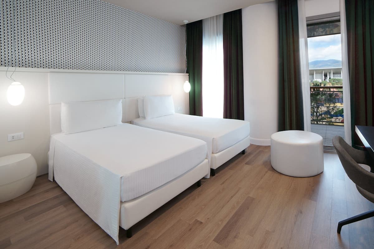 A modern hotel room with two beds, a round ottoman, a desk, and a large window with a view.