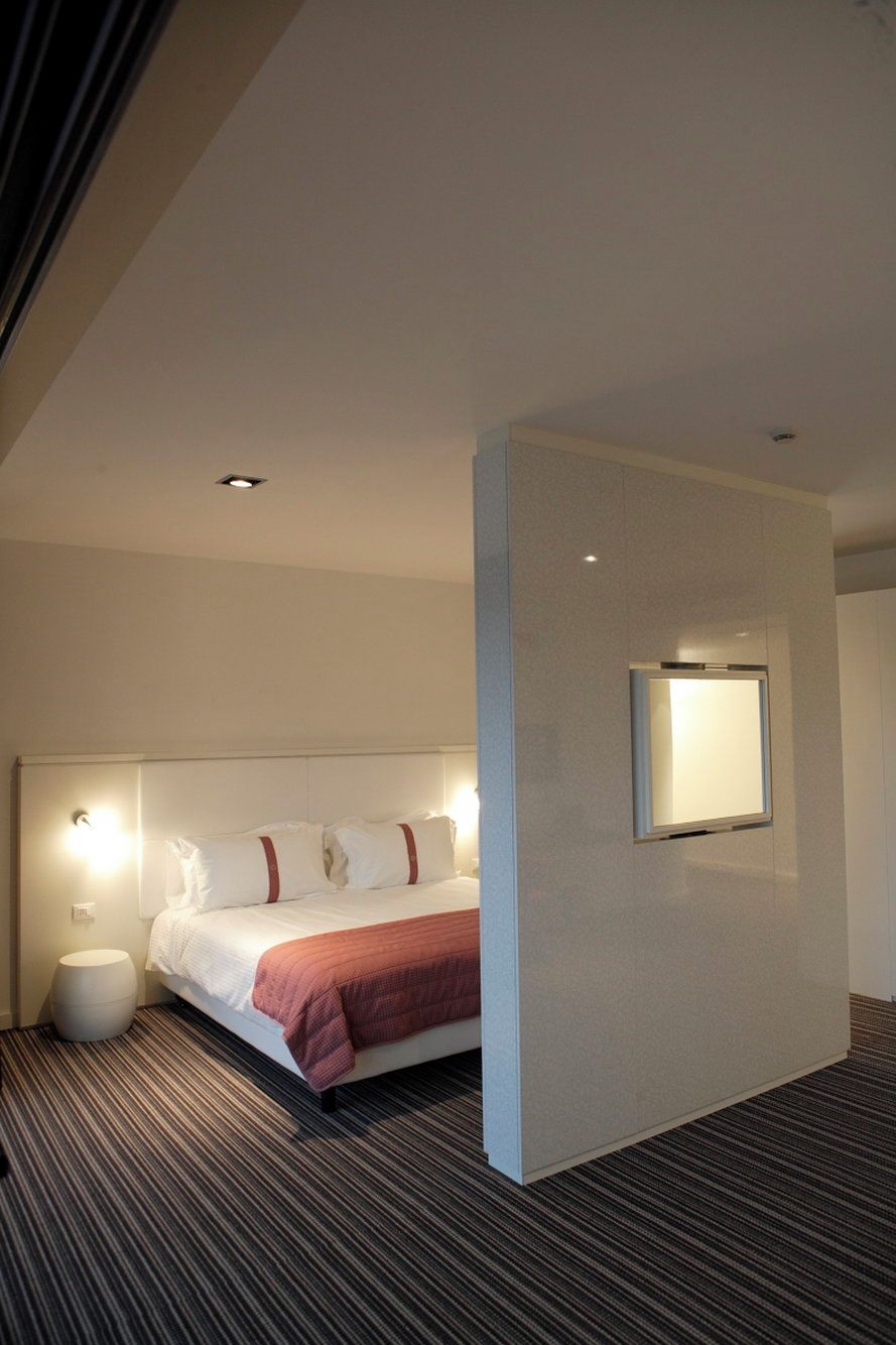 A modern hotel room with a bed, a bedside table, a wall-mounted light, and a carpeted floor.