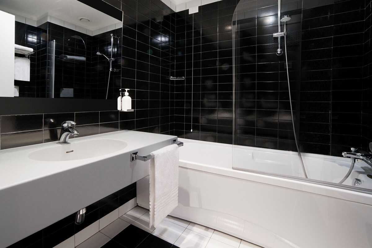 Ein modernes Badezimmer mit einem weißen Waschbecken, einem Spiegel, einer Badewanne und schwarzen Fliesenwänden.