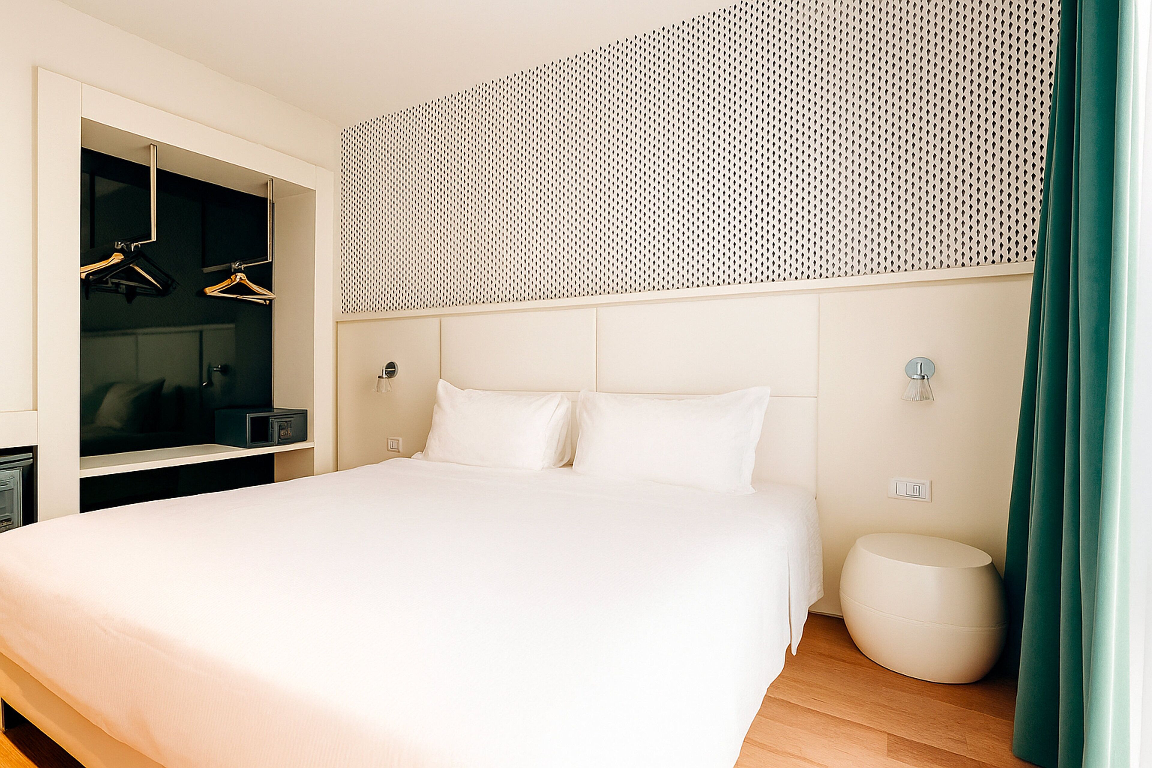 A modern hotel room with a large bed, a walk-in closet with hanging clothes, a round ottoman, and a patterned wall.