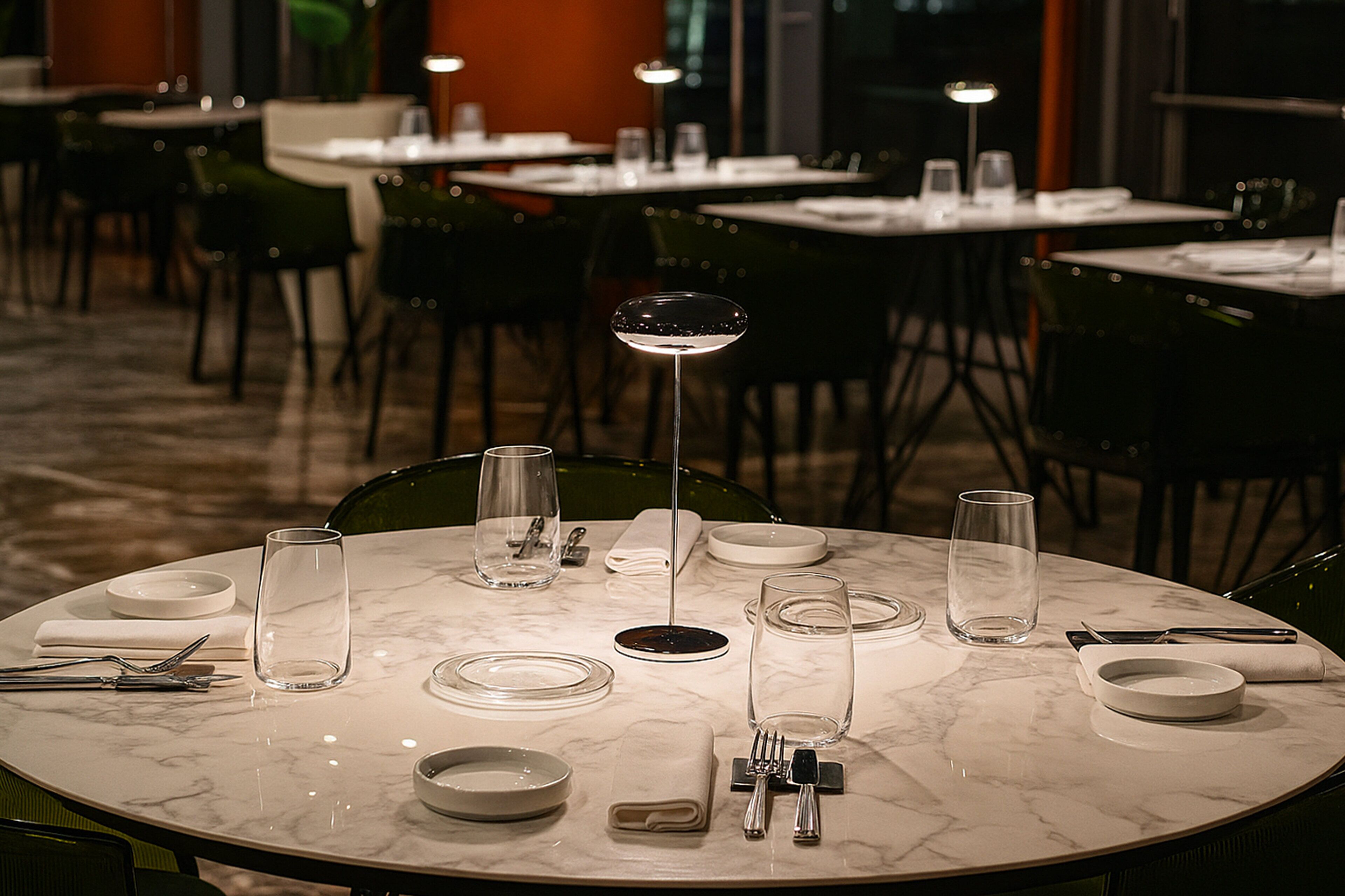 A dining area with round tables set for four, each with a wine glass, plates, cutlery, and a folded napkin.