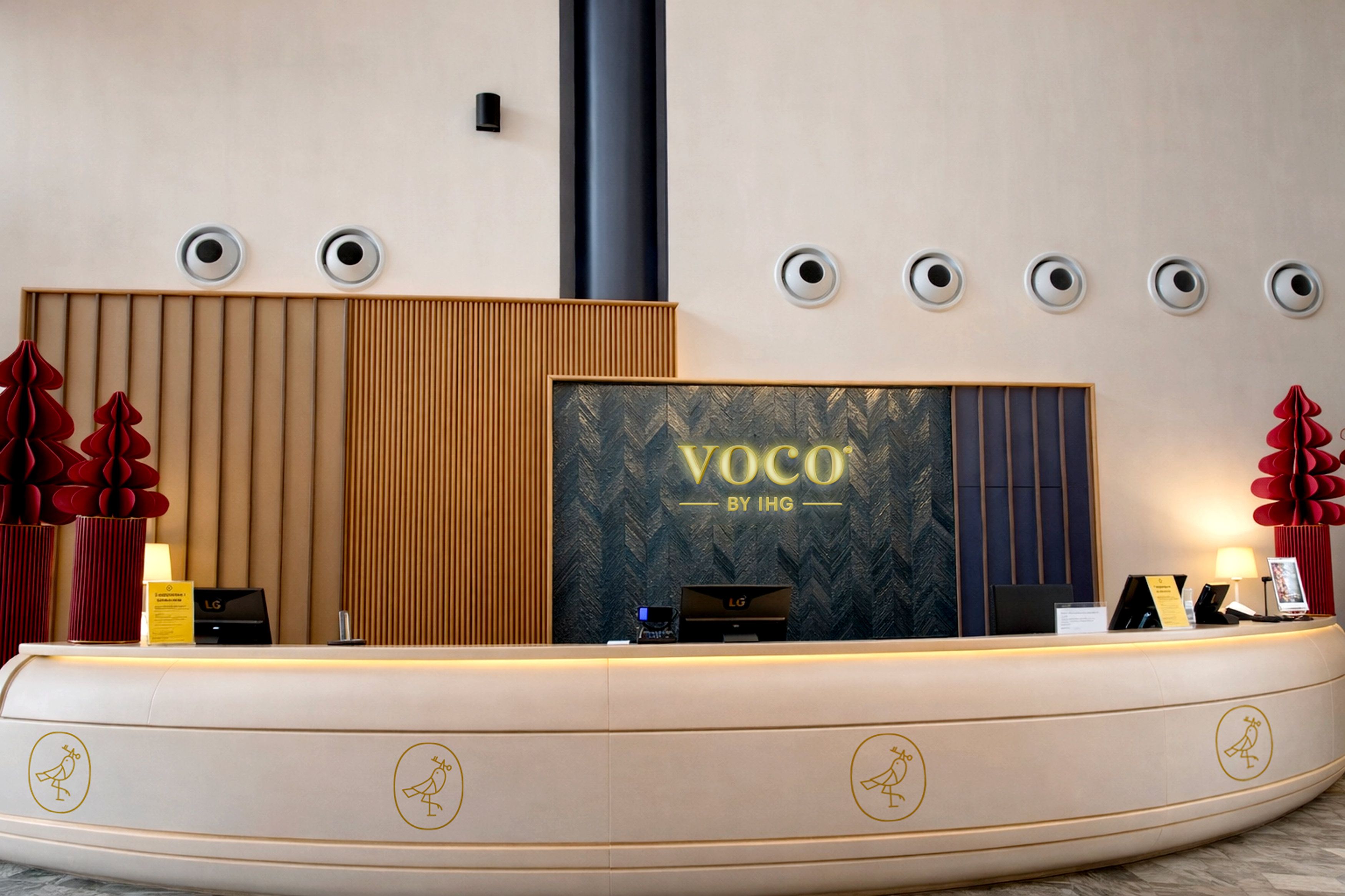 A reception desk with a large screen displaying 'VOCO BY IHG', a modern design with vertical wooden panels, and decorative red vases.