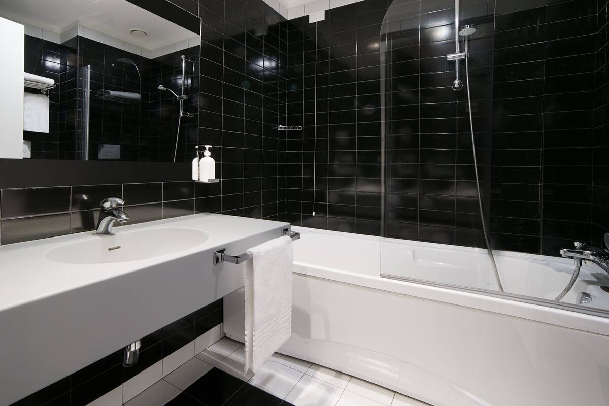 A modern bathroom with a white sink, a mirror, a bathtub, and black tiled walls.