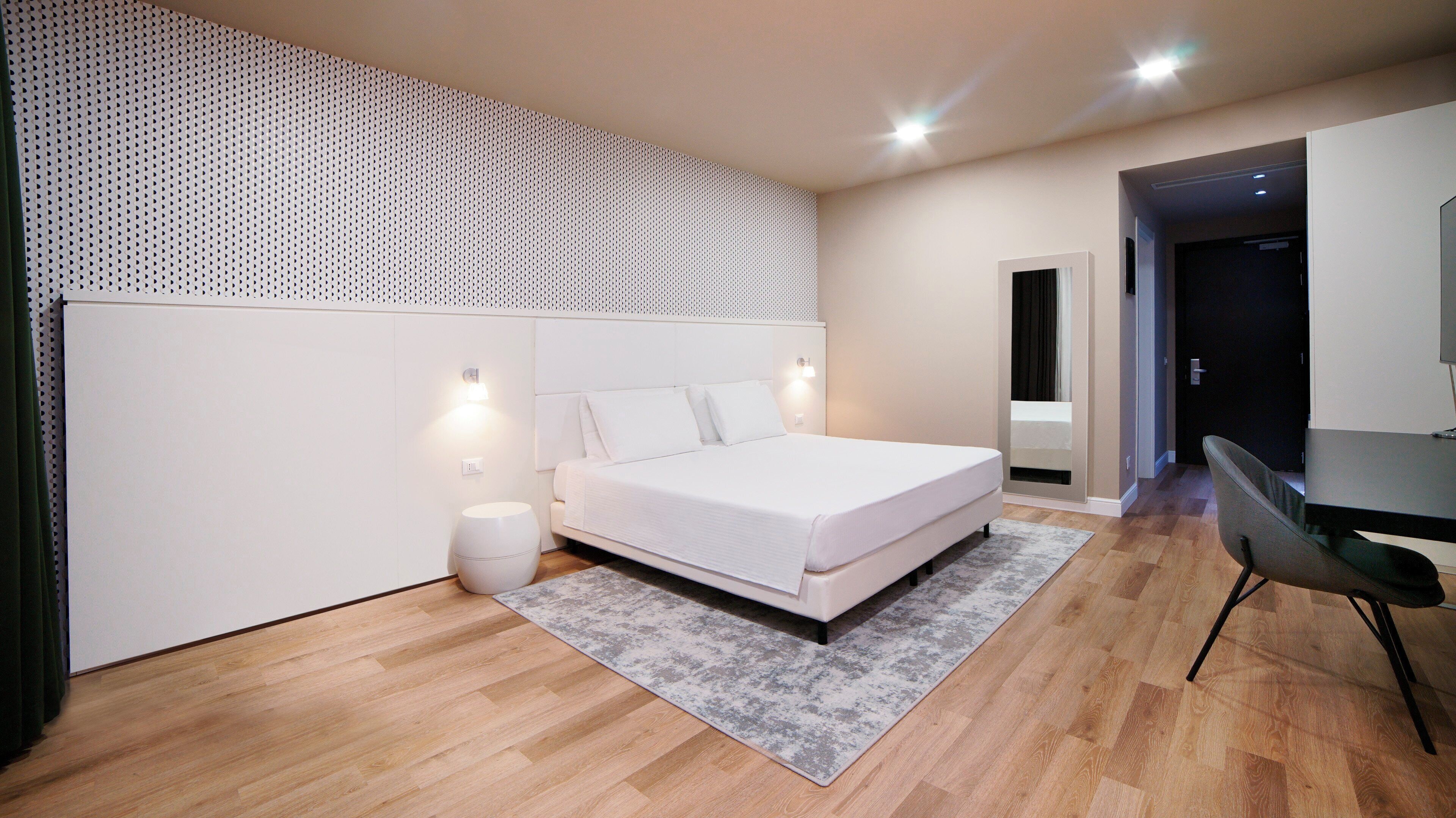 Un dormitorio moderno con una cama grande, un taburete pequeño y redondo, una alfombra gris y suelo de madera.