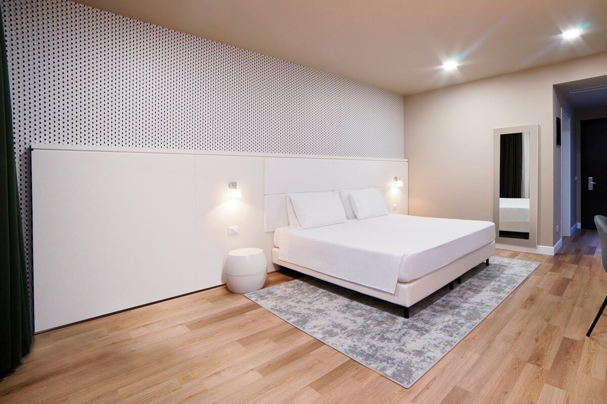 A modern bedroom with a large bed, a small round stool, a grey rug, and a wooden floor.