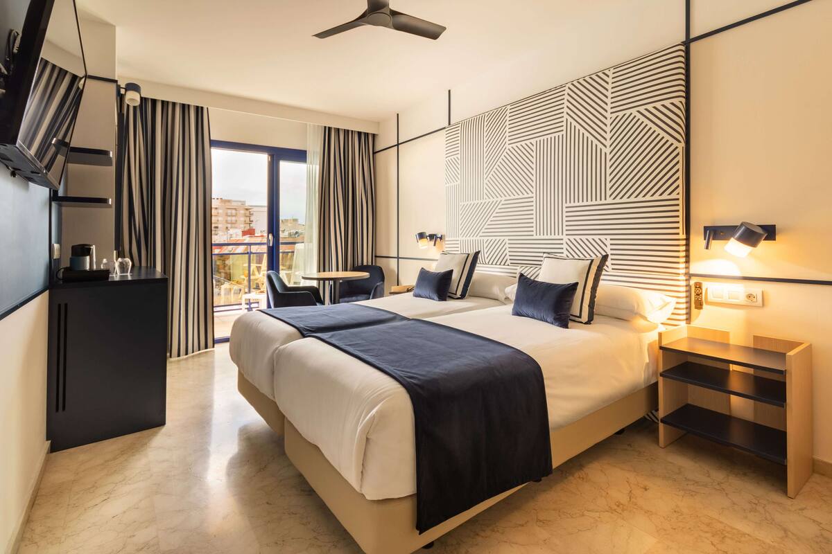 A modern hotel room with a large bed, a desk, a chair, a TV, and a balcony with curtains.