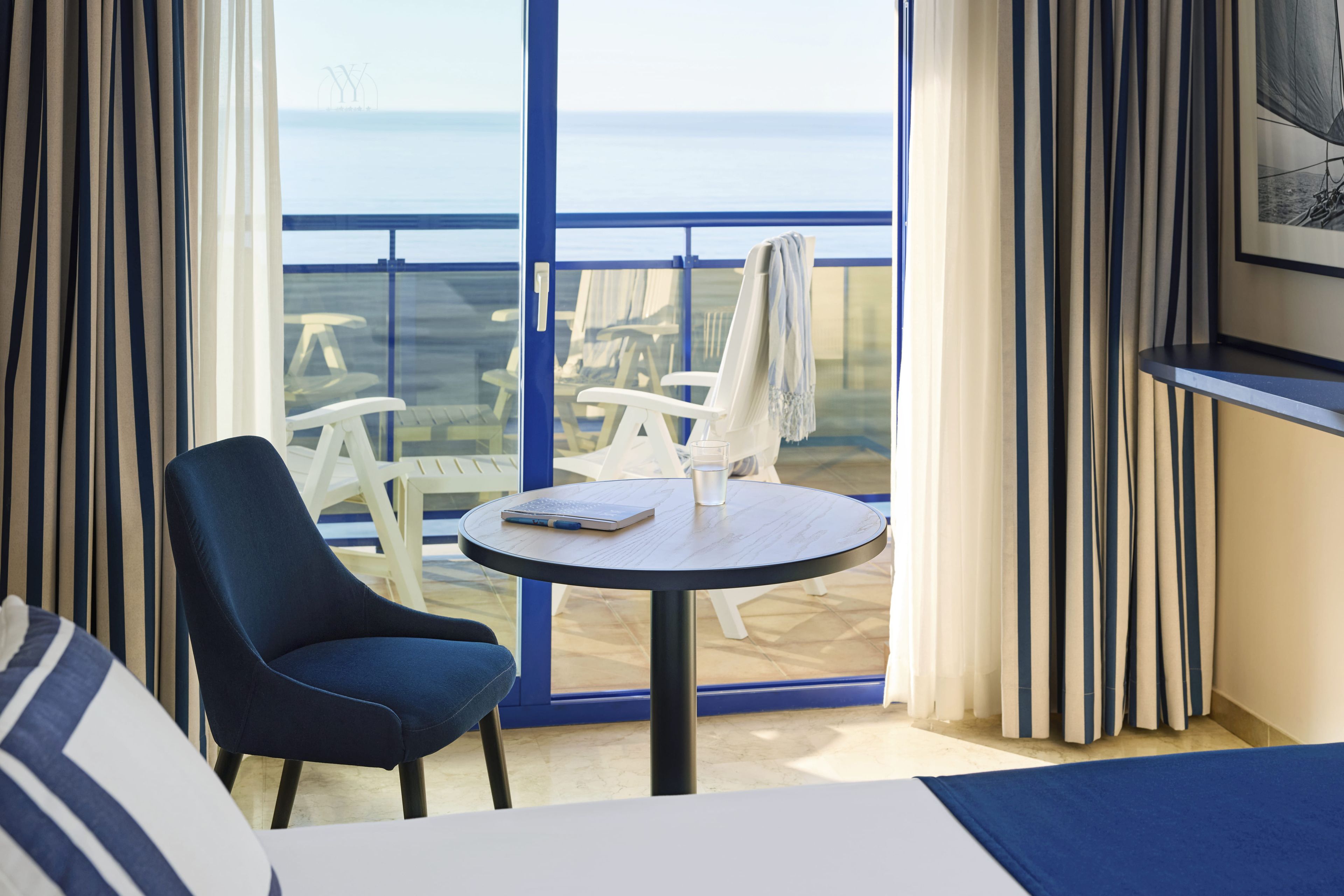 Habitación de hotel con cama, una mesa redonda con una laptop y vista al mar a través de amplias ventanas.