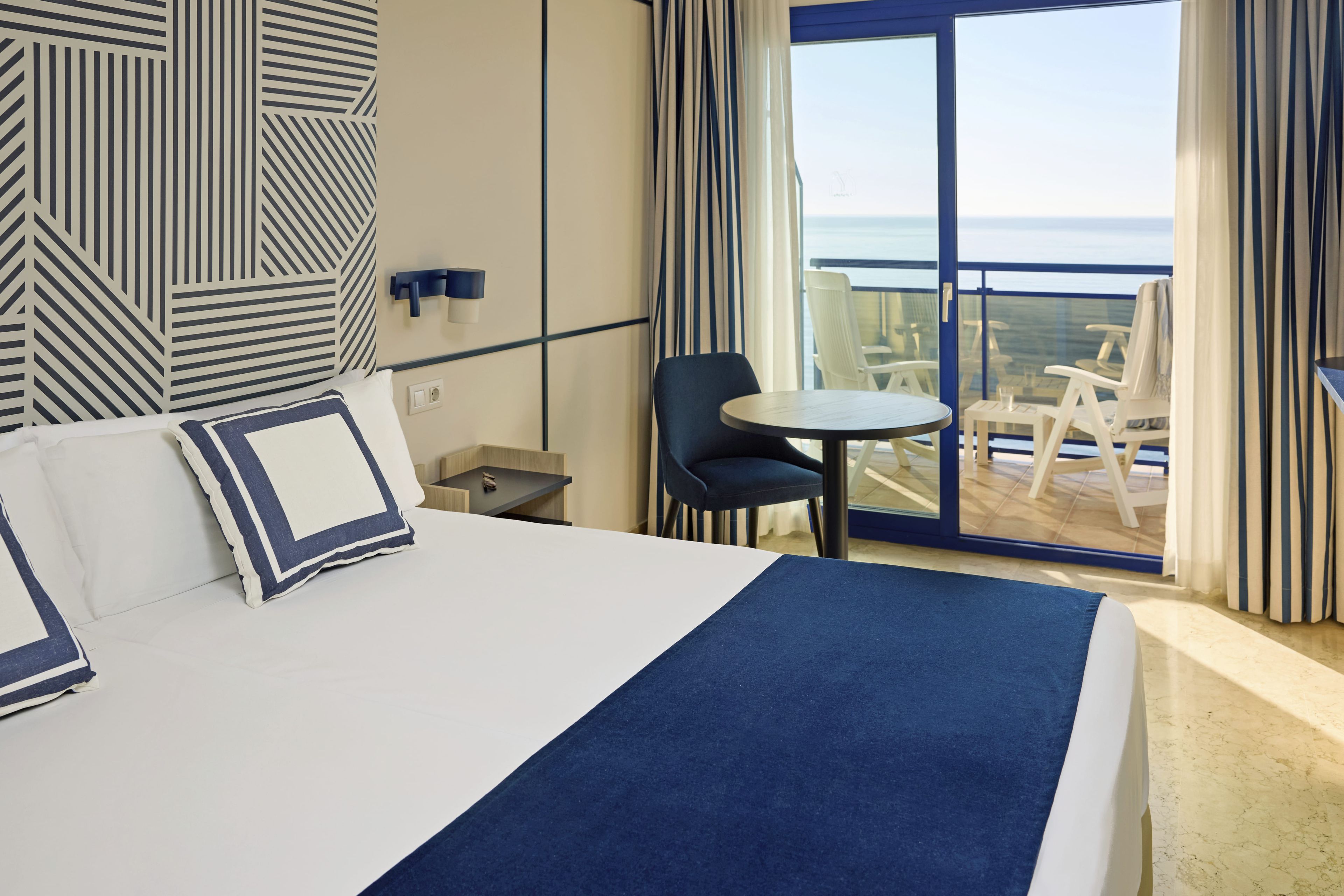 A hotel room with a bed, a small table, a chair, and a balcony with a view of the sea.