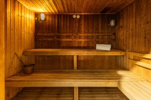 A wooden sauna with benches and a bench seat, a metal bucket, and wall-mounted clocks.