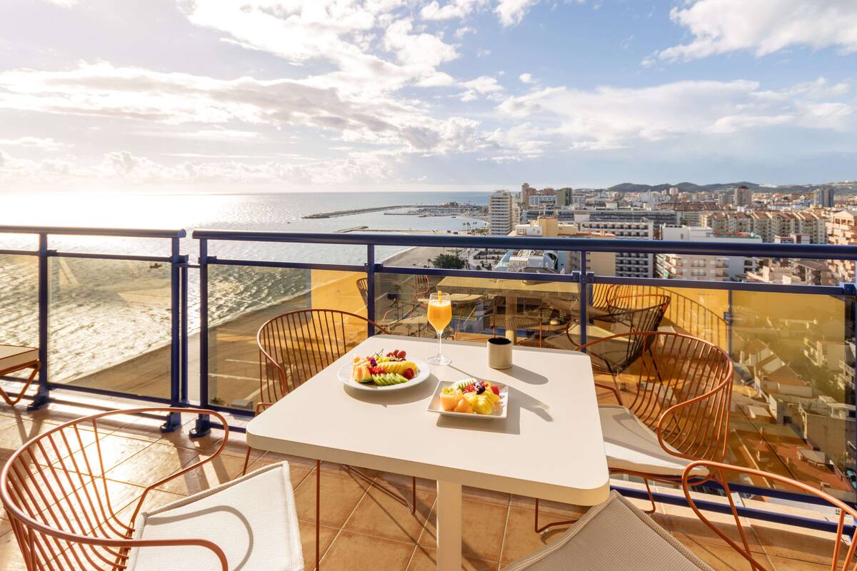 A balcony with a table set for two, featuring a plate of fruit and a glass of orange juice. The balcony overlooks a beach and cityscape.