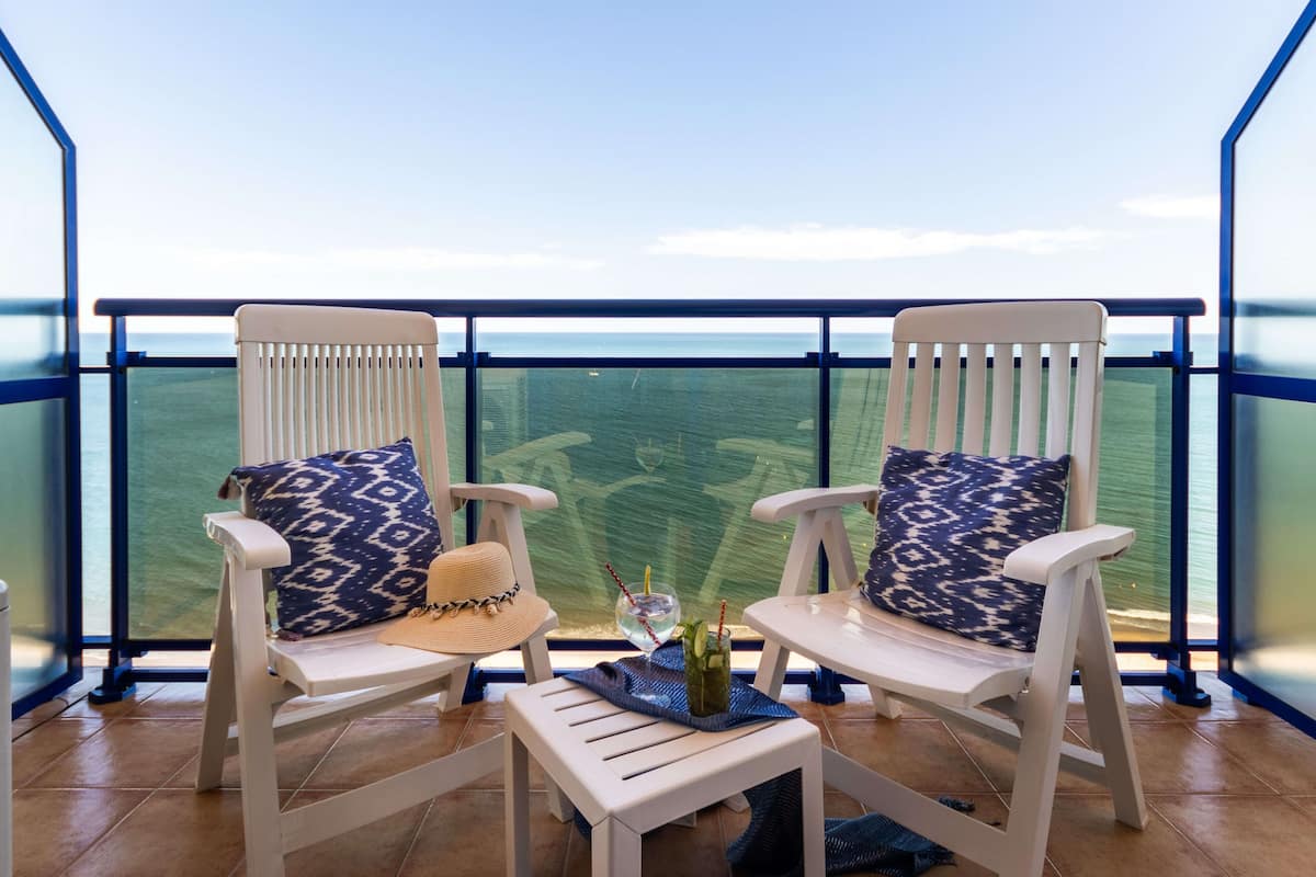 Deux chaises blanches avec des coussins à motifs, une petite table et un panier en osier sur un balcon donnant sur la mer.