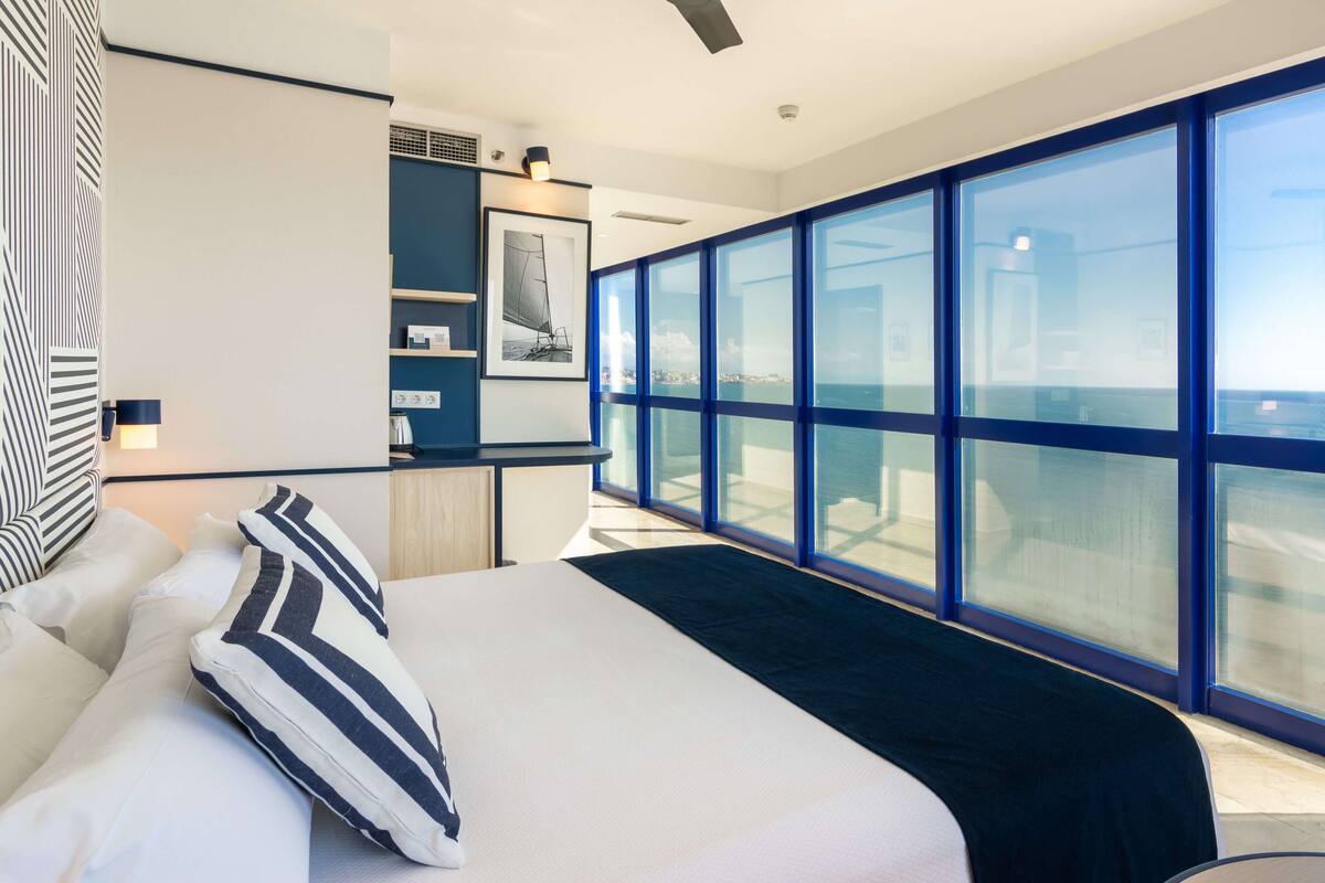 A modern hotel room with a large bed, a desk, a view of the sea, and a balcony.