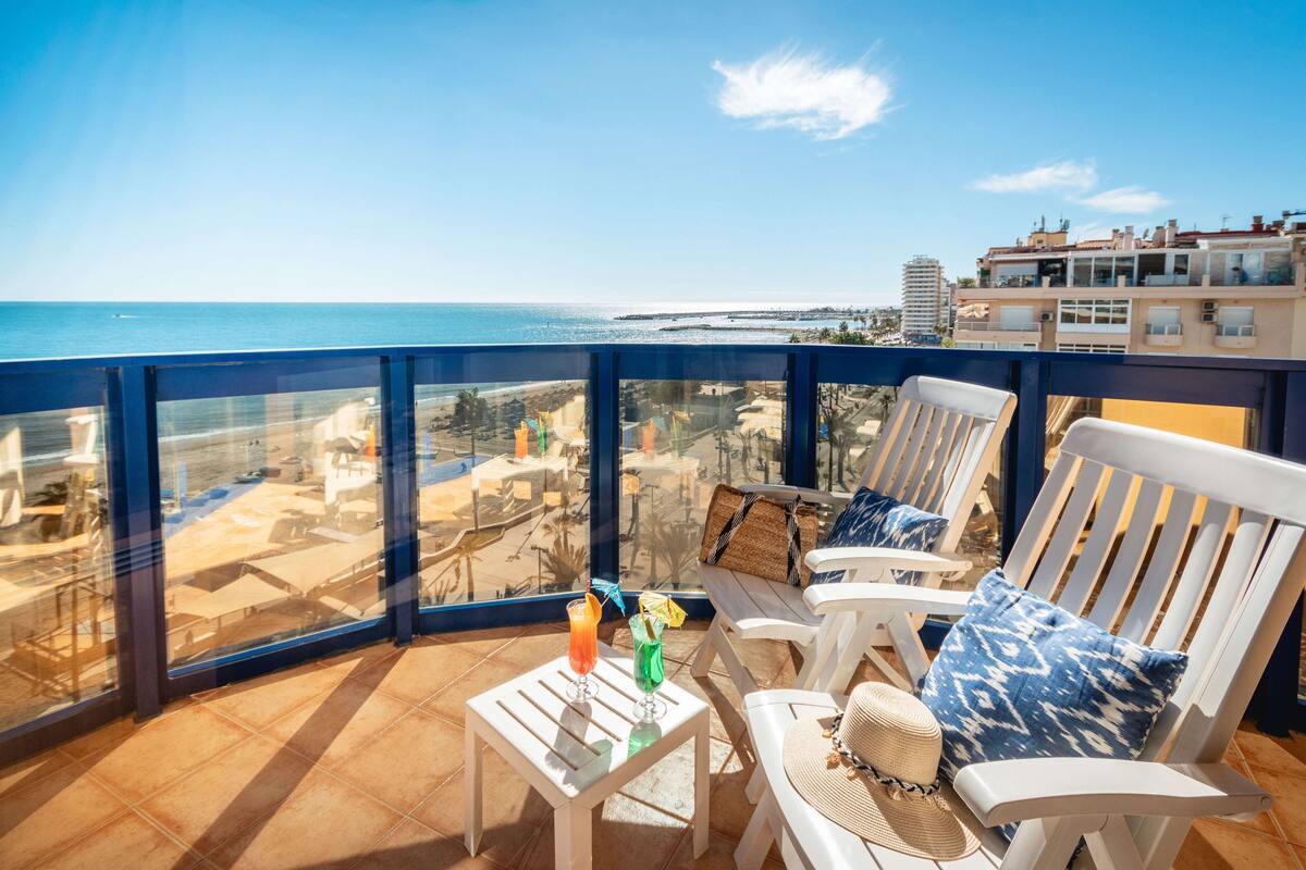 A balcony with a view of the beach, white chairs, a small table with drinks, and a woven basket.