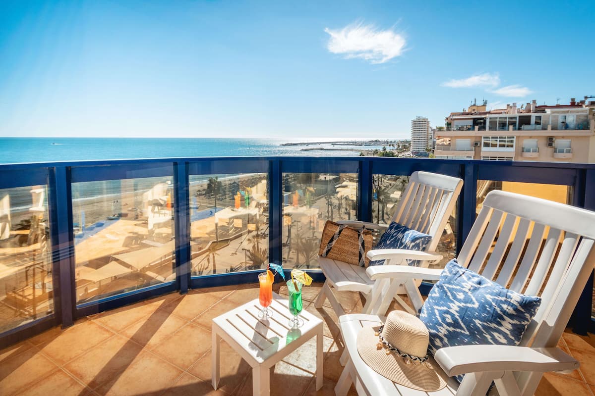 Un balcon donnant sur la plage, des chaises blanches, une petite table avec des boissons et un panier en osier.