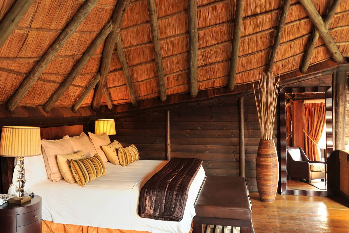 Una habitación de cabaña de madera con una cama, lámparas de noche, un jarrón grande y un banco de madera.