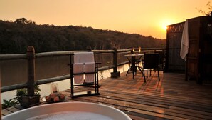 A wooden deck with a table and chairs, overlooking a body of water and a forested hillside during sunset.