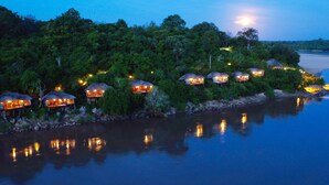 A series of thatched-roof huts along a riverside, surrounded by dense greenery.