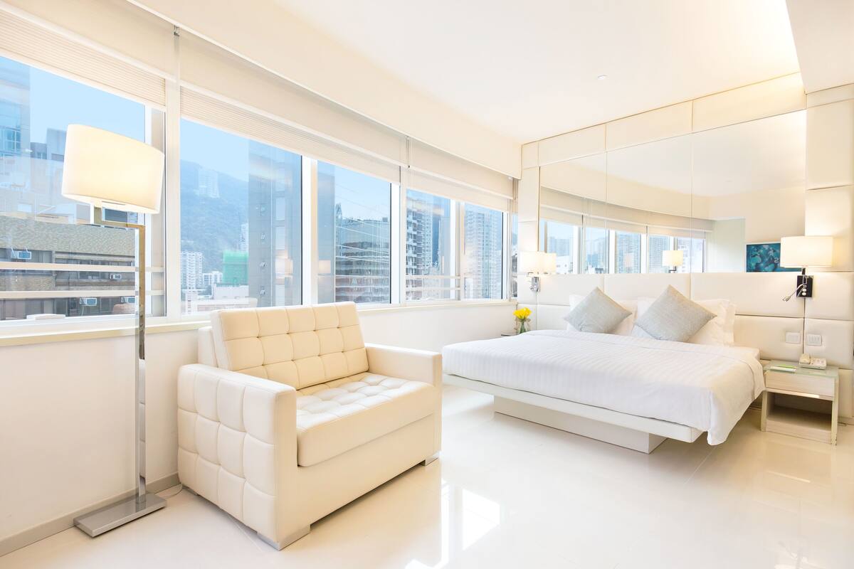 Ein modernes Hotelzimmer mit einem großen Bett, einer Couch, einer Stehlampe und Blick auf die Stadt durch große Fenster.