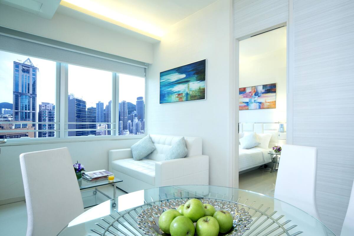 Ein modernes Wohnzimmer mit einem Glastisch, weißen Sofas und Blick auf die Stadt durch große Fenster.