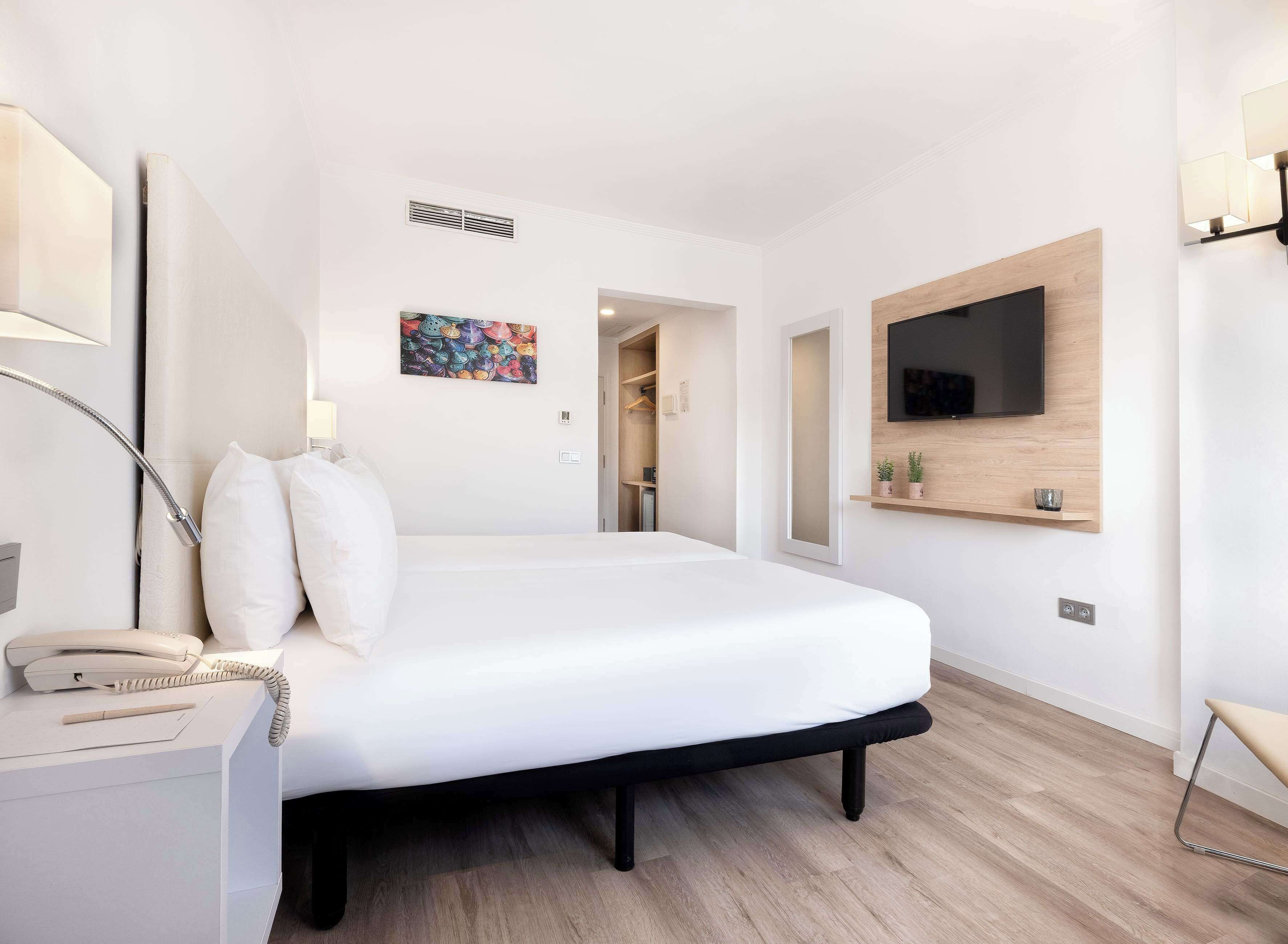 A modern hotel room with a large bed, a bedside telephone, a desk with a lamp, a flat-screen TV mounted on the wall, and a small shelf with decorative items.