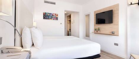 A modern hotel room with a large bed, a bedside telephone, a desk with a lamp, a flat-screen TV mounted on the wall, and a small shelf with decorative items.