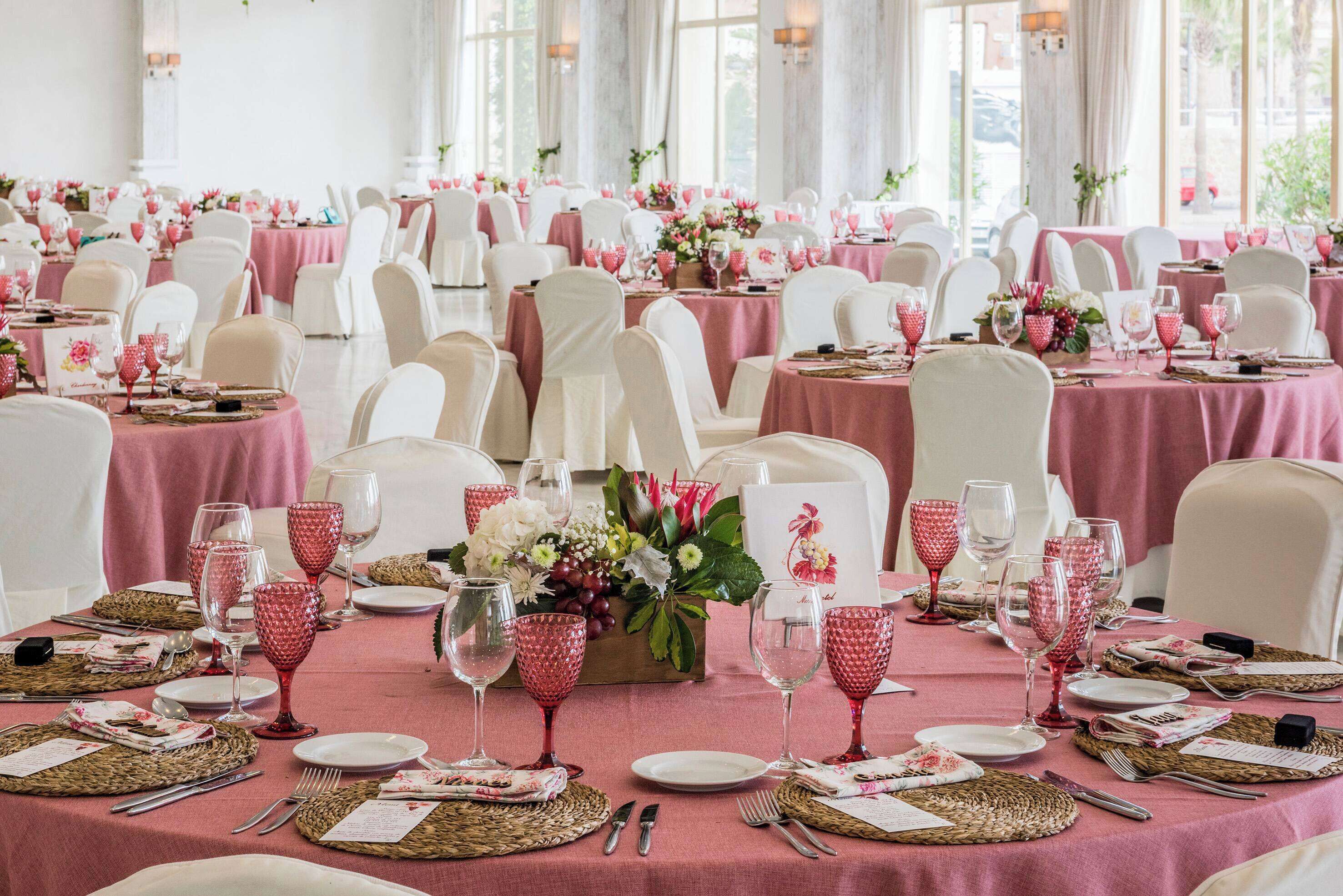 A banquet hall with tables set for a formal event, featuring pink tablecloths, white chairs, and red glassware.