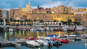 A marina with boats docked, a historic building with a tower, and a waterfront promenade. - Hotel Melilla Puerto (Melilla)
