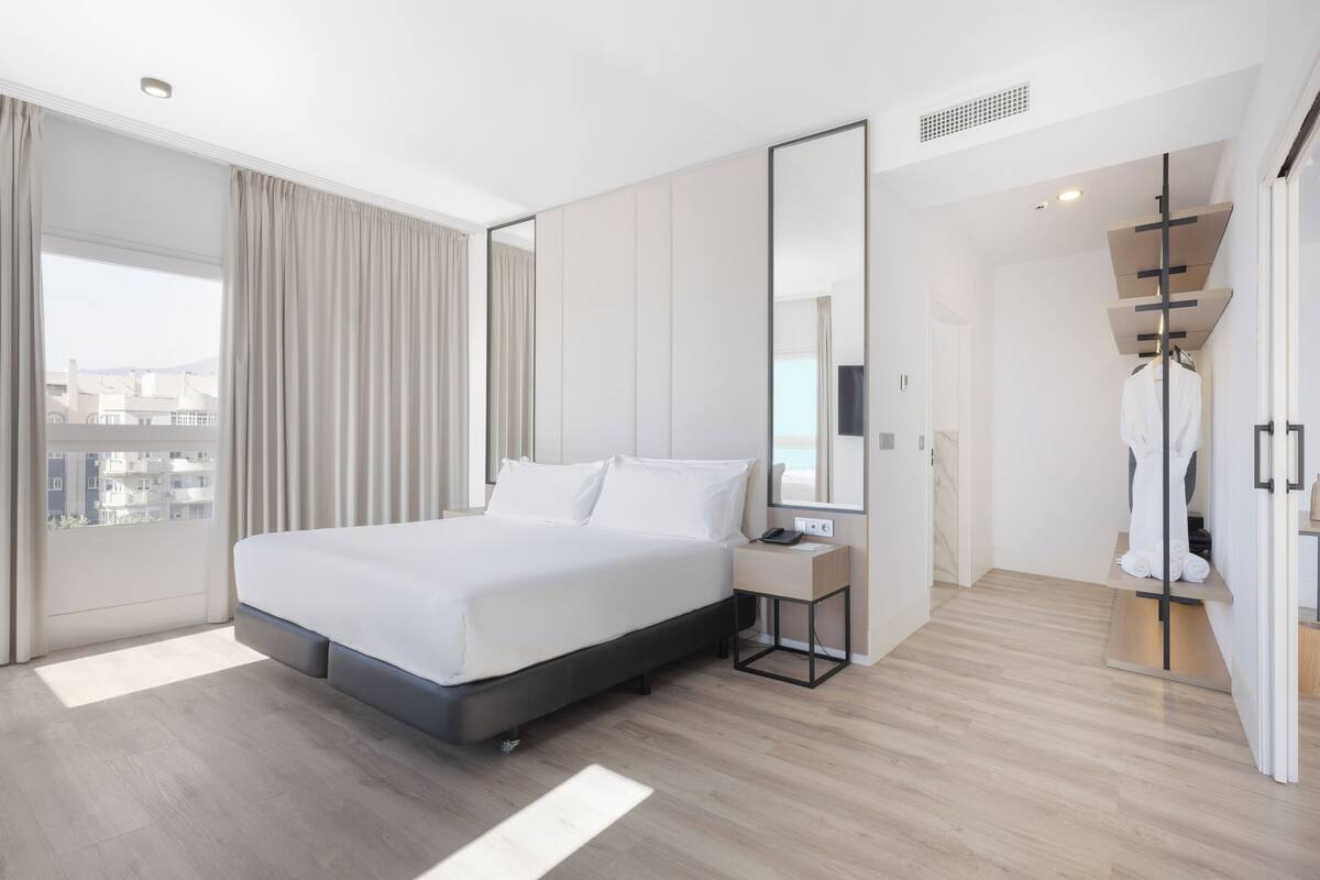 Ett modernt hotellrum med en stor säng, ett sängbord med en telefon, en spegel och utsikt över staden genom stora fönster.
