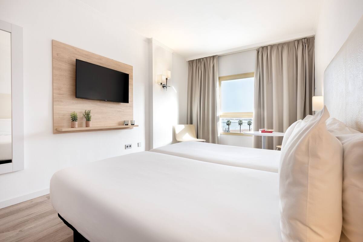 Ett hotellrum med en stor säng, en tv som är monterad på en träpanel, ett fönster med gardiner och utsikt över palmer.