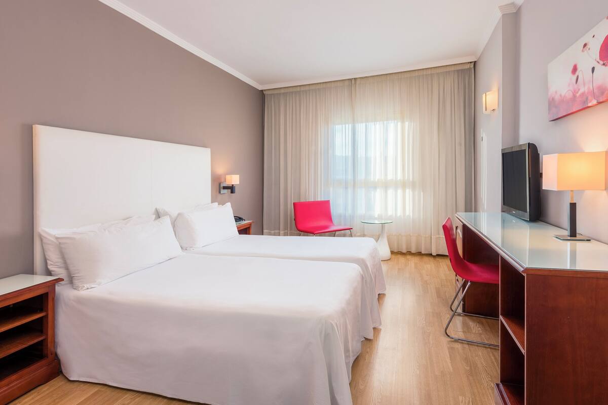 Ett hotellrum med två sängar, ett skrivbord, en stol, en TV och ett fönster med gardiner.