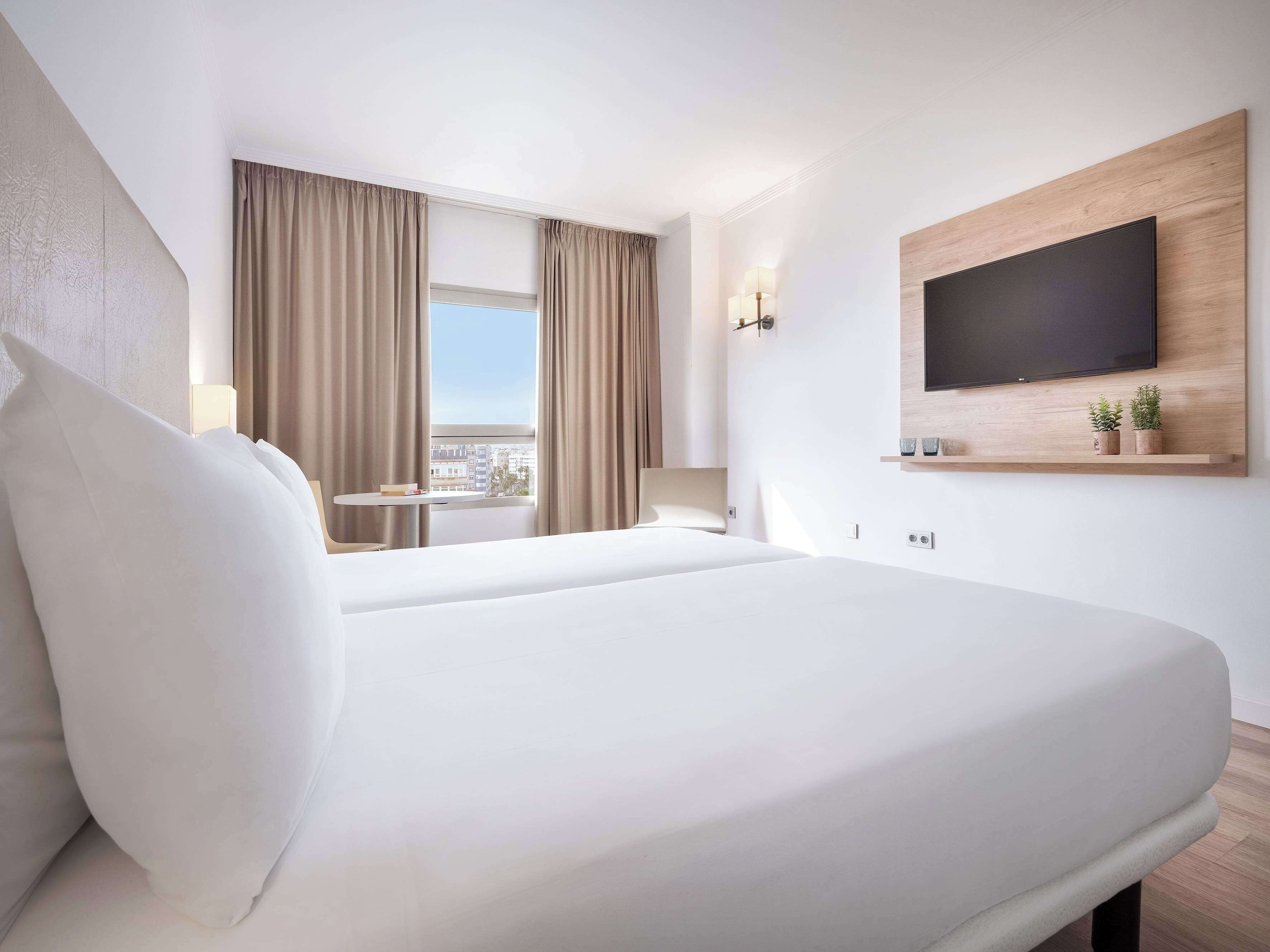 Habitación de hotel con una cama grande, un televisor en la pared, una mesita y vistas a la ciudad desde la ventana.