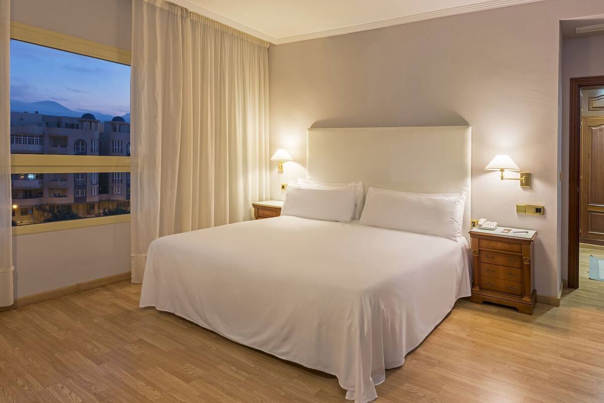 Ett hotellrum med en stor säng, två sänglampor, ett nattduksbord, ett fönster med gardiner och utsikt över byggnader utanför.