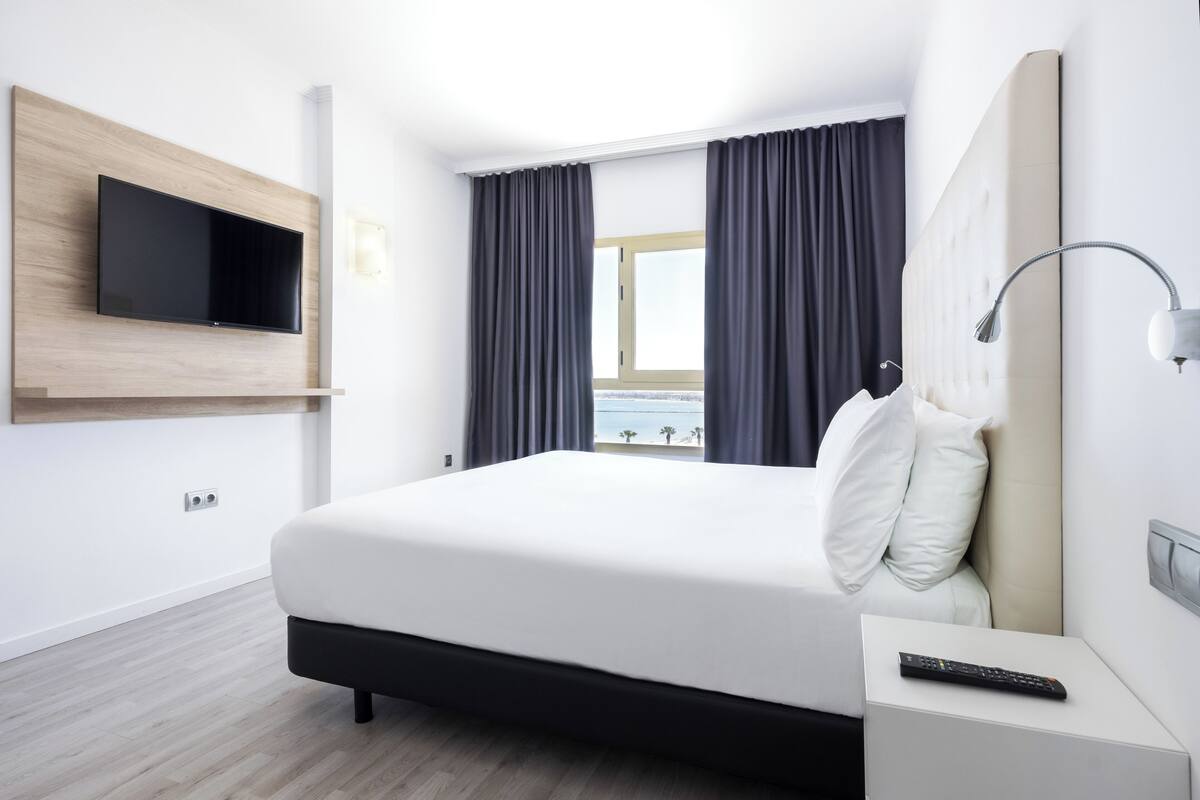 Ett modernt hotellrum med en stor säng, en väggmonterad TV, en sänglampa och utsikt över omgivningarna genom fönstret.
