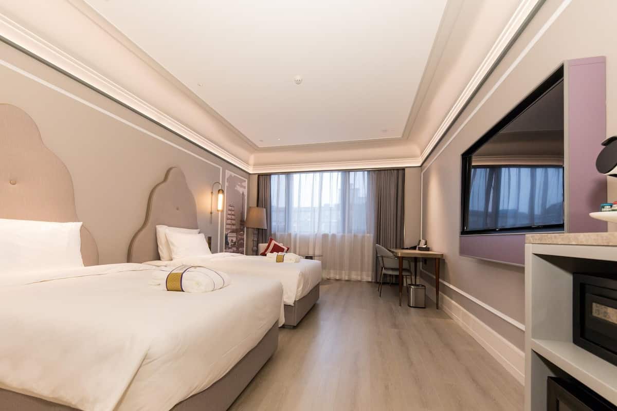 A hotel room with two beds, a desk, a TV, and a window with curtains.
