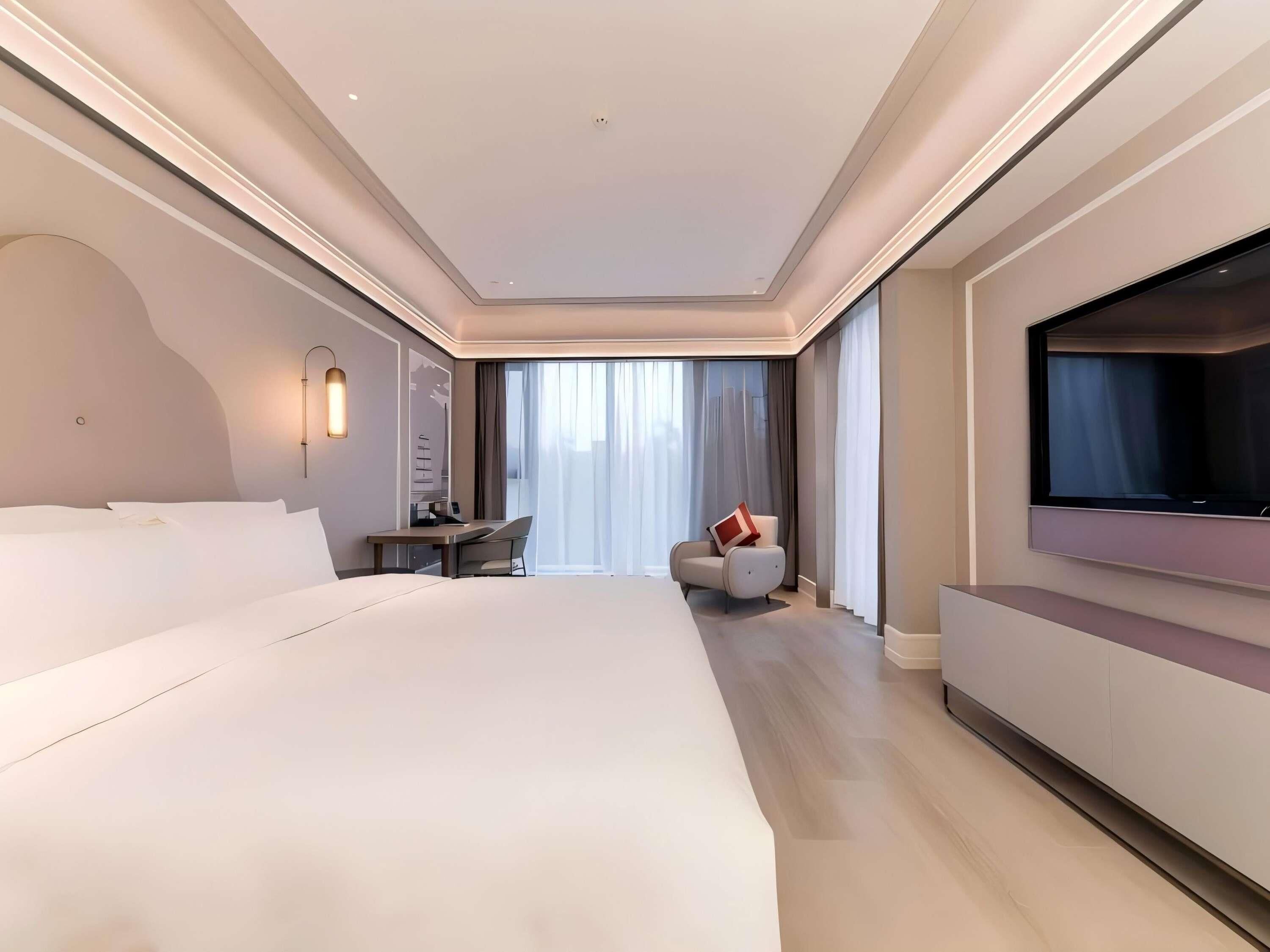 Quarto de hotel moderno com uma cama grande, uma mesa de trabalho, uma cadeira, televisão e uma janela ampla com cortinas.
