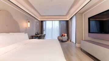 Quarto de hotel moderno com uma cama grande, uma mesa de trabalho, uma cadeira, televisão e uma janela ampla com cortinas.