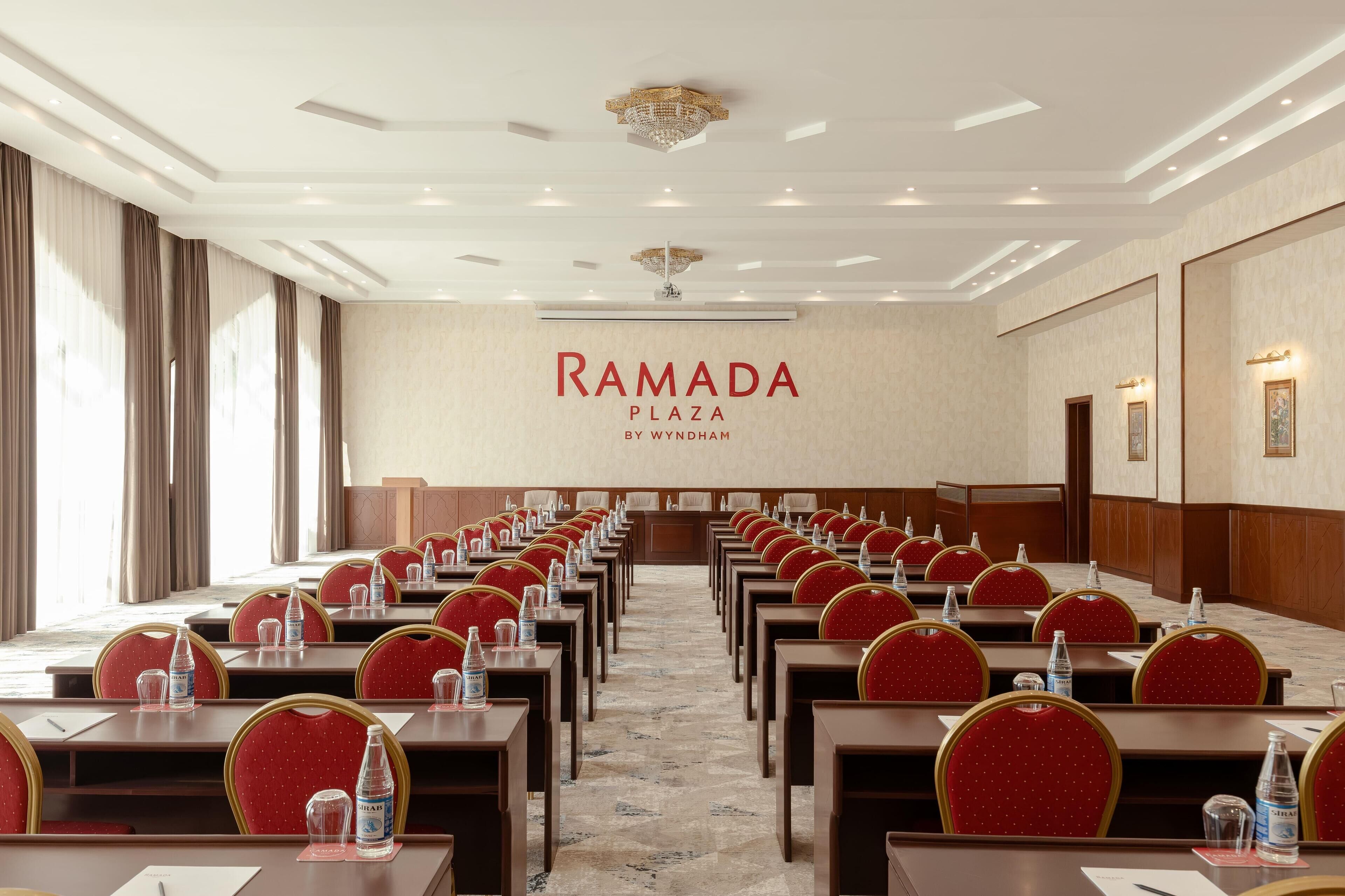 A conference room with rows of tables and chairs, a chandelier, and a wall with the text "RAMADA PLAZA BY WYNDHAM".