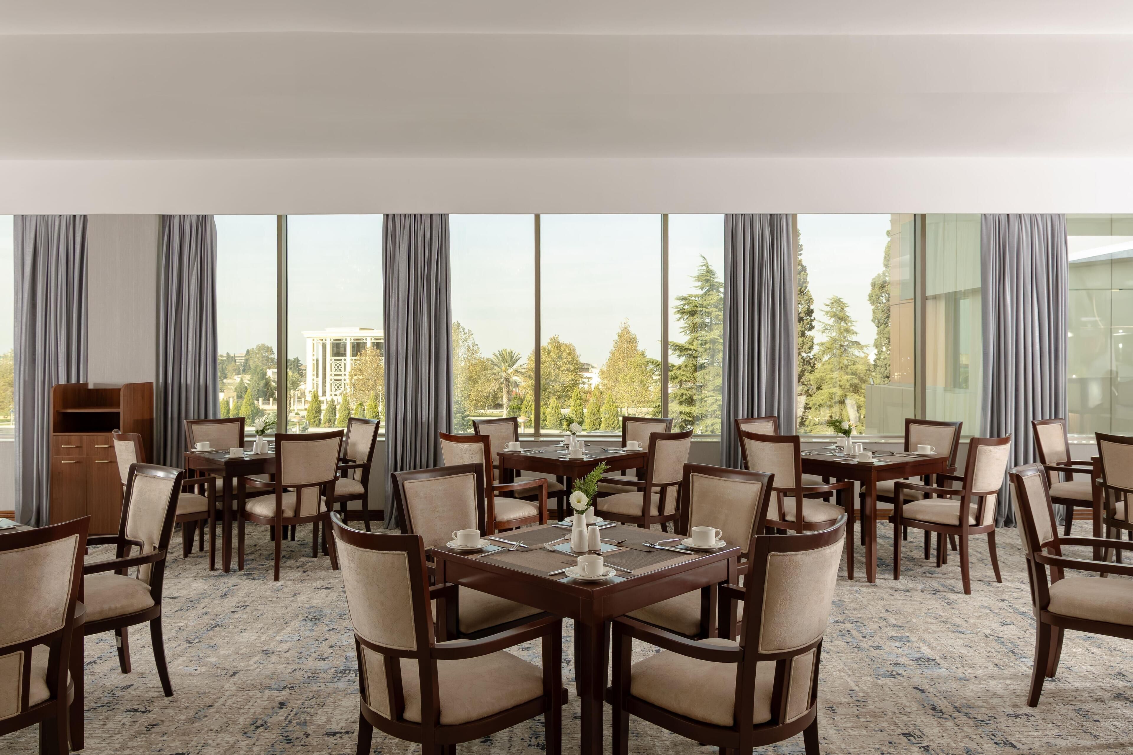 Een restaurant in een hotel met meerdere tafels en stoelen, grote ramen met gordijnen, en uitzicht op bomen en gebouwen.