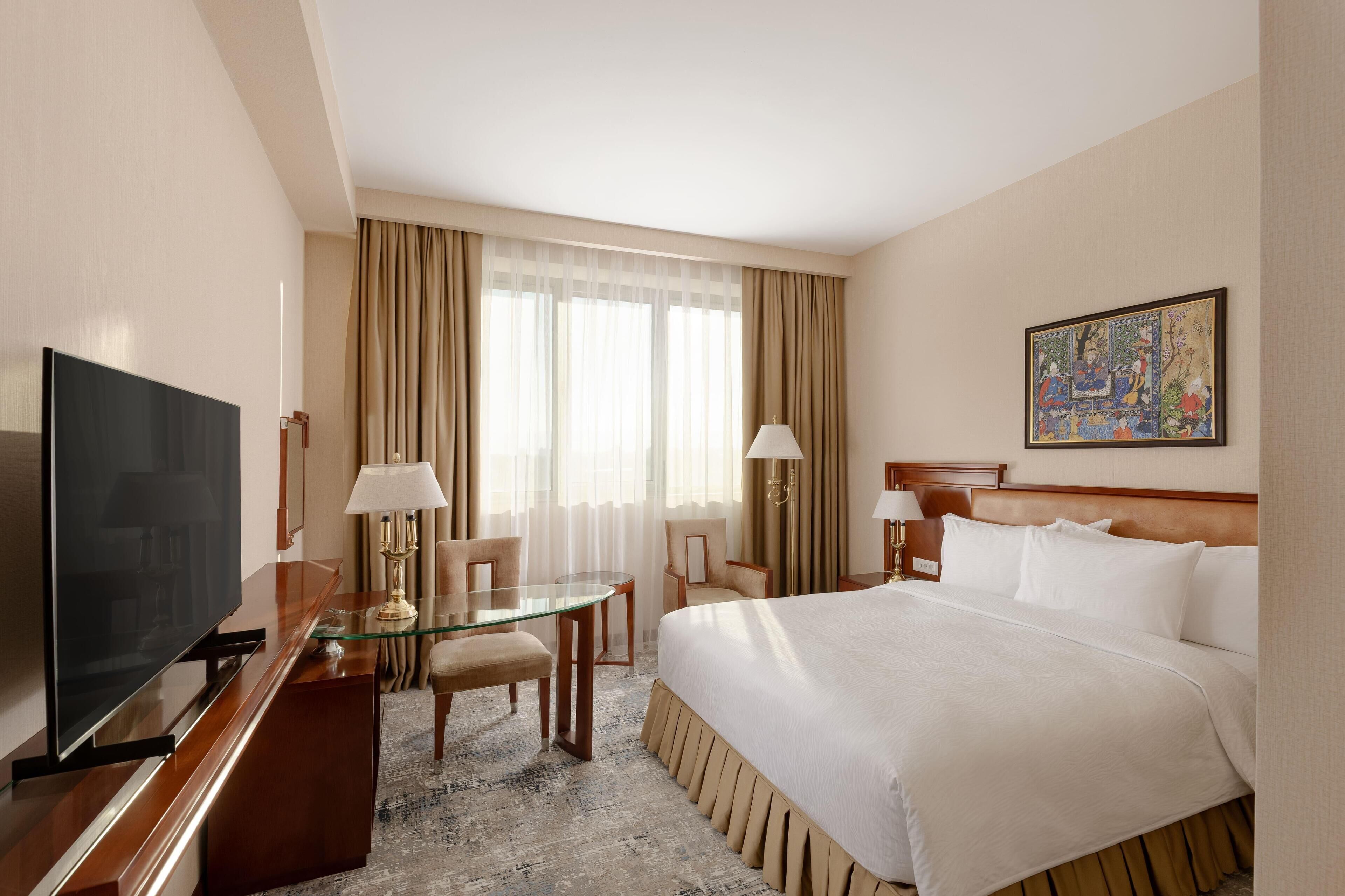 A hotel room with a large bed, a TV, a glass table, two chairs, a desk, a lamp, a painting on the wall, and a window with curtains.